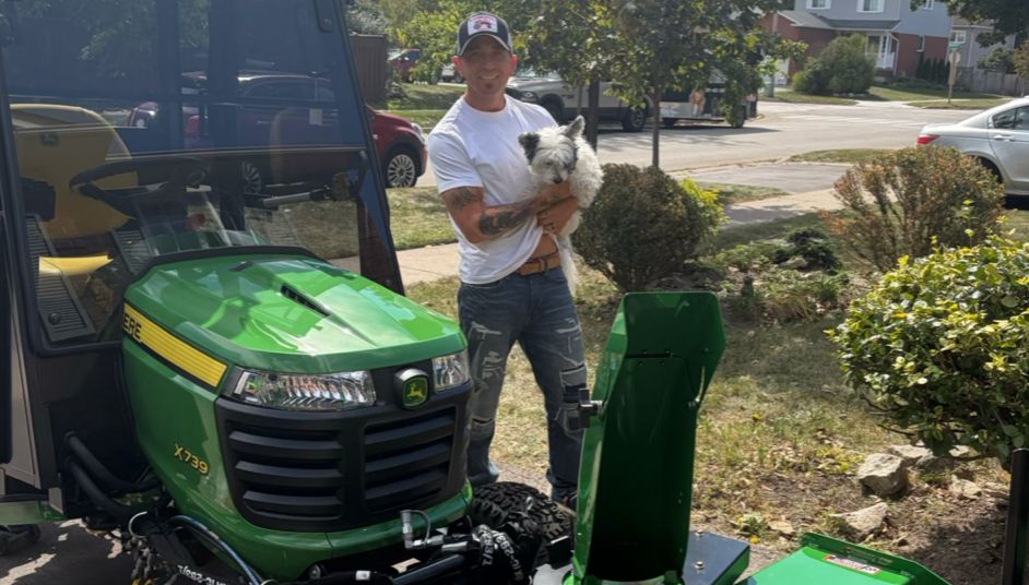 A man holding a small white dog beside the green cab tractor in a front yard